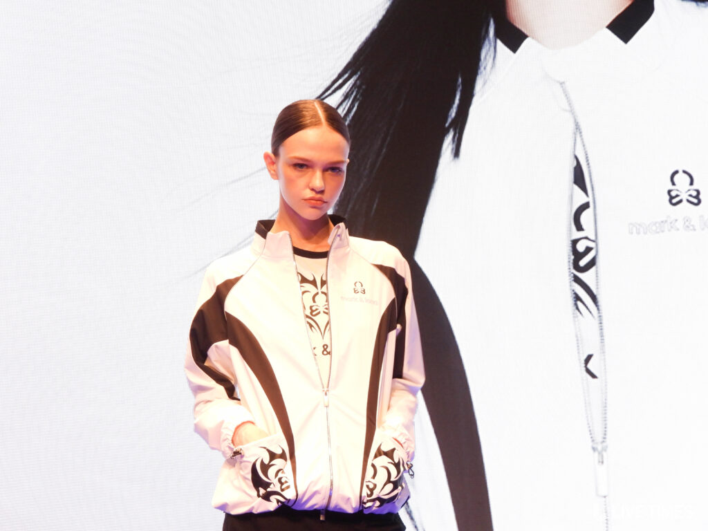 Fashion model wearing a white and black patterned jacket on a runway, hands in pockets, serious expression.