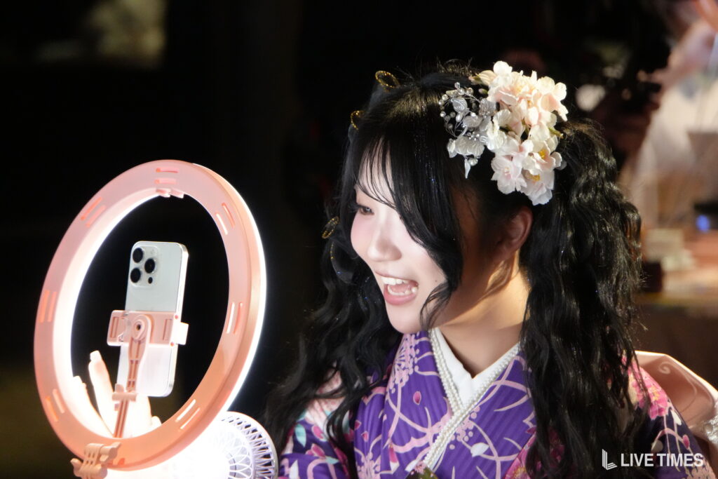 Woman wearing a floral headpiece smiles at a smartphone mounted in a pink ring light for a selfie photo.