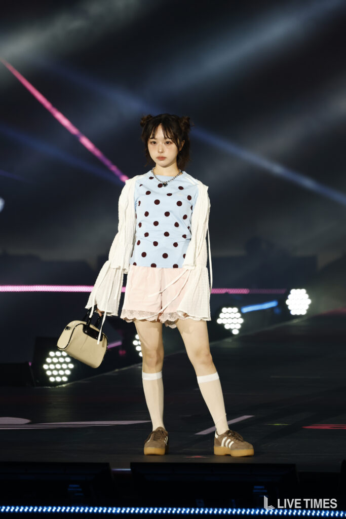 Stage performer on a dark backdrop wearing a light blue polka-dot top, white cardigan, pink skirt, knee-high socks, and a beige handbag under bright lights.