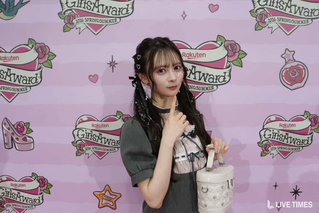 Young woman posing at a GirlsAward event, wearing a gray dress and holding a white handbag in front of a pink backdrop with logos.