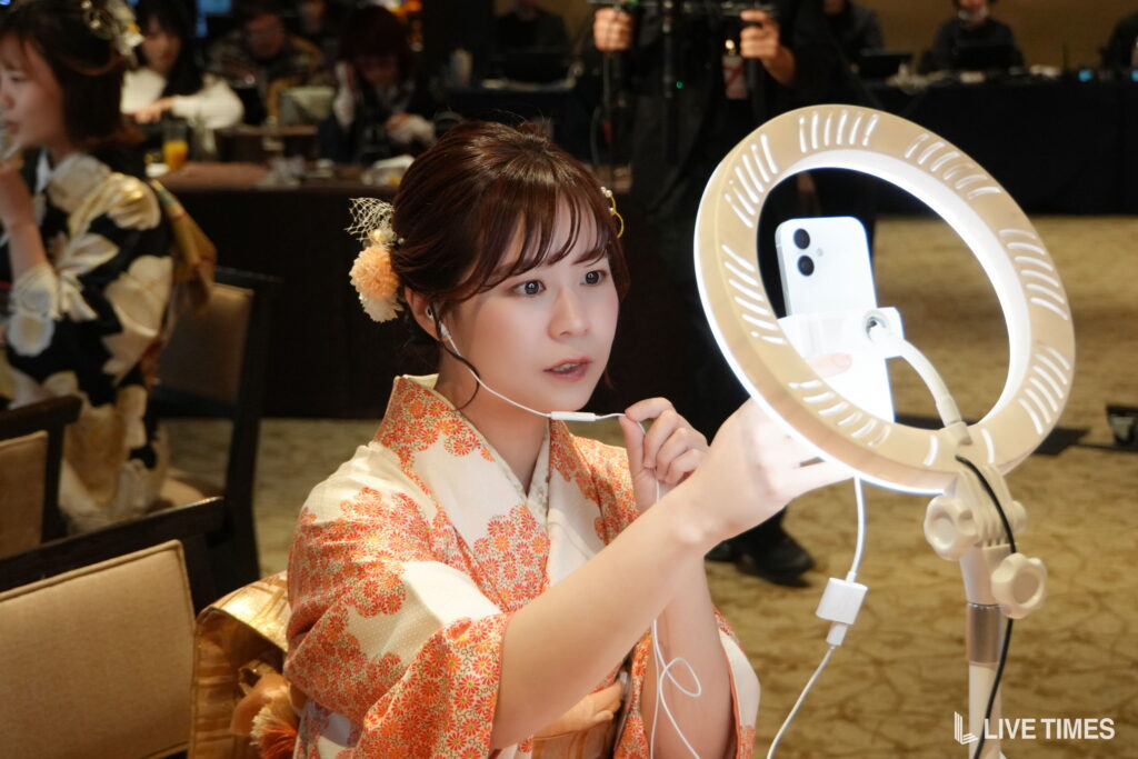 Woman in a floral kimono adjusts a ring light and a smartphone mounted on a stand during an event, with people seated in the background.