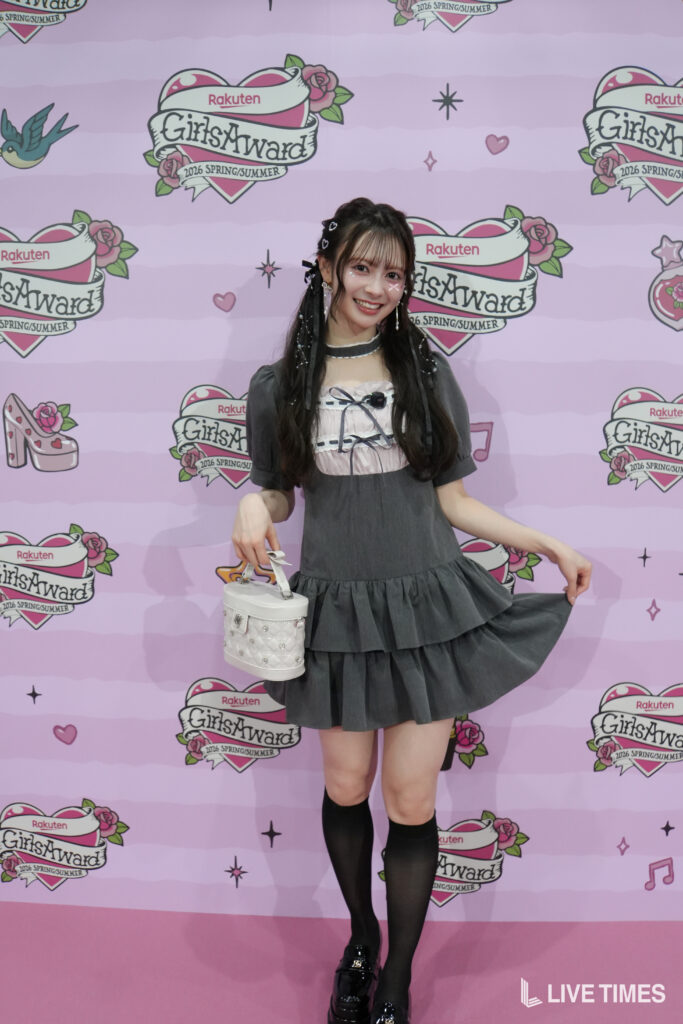 Smiling young woman in a gray layered dress, black knee-high socks, and a white handbag posing in front of a pink GirlsAward backdrop.