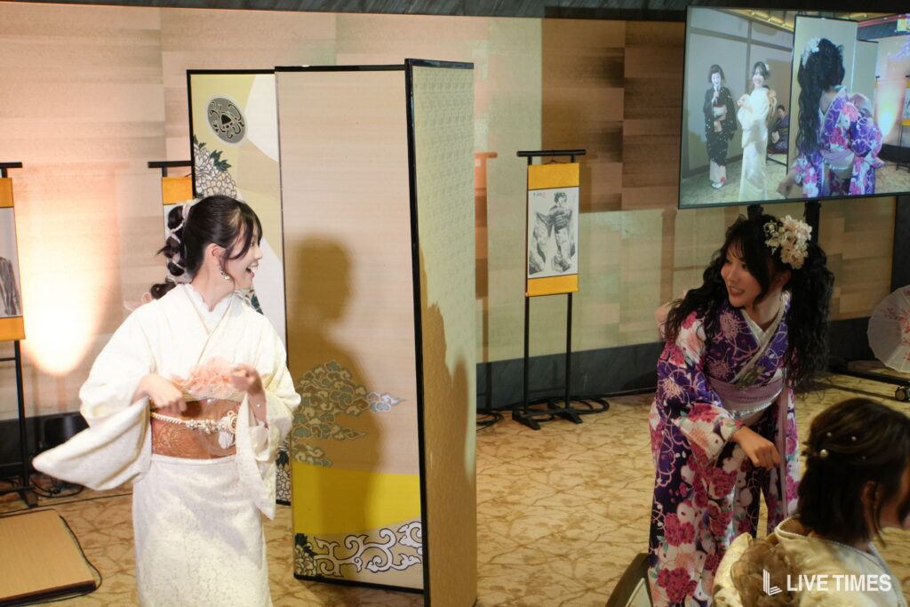 Two women in colorful kimonos smile and chat indoors beside decorative folding screens at an event.
