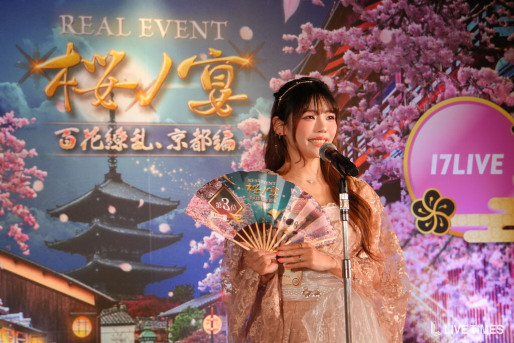 Smiling woman in a pink dress speaks into a microphone on a stage with cherry blossom backdrop and event signage behind her.
