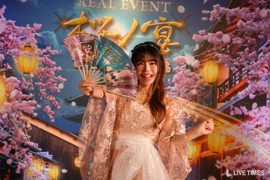 Smiling young woman in a lace dress holds decorative fans in front of a cherry-blossom festival backdrop with lanterns and pink blossoms.