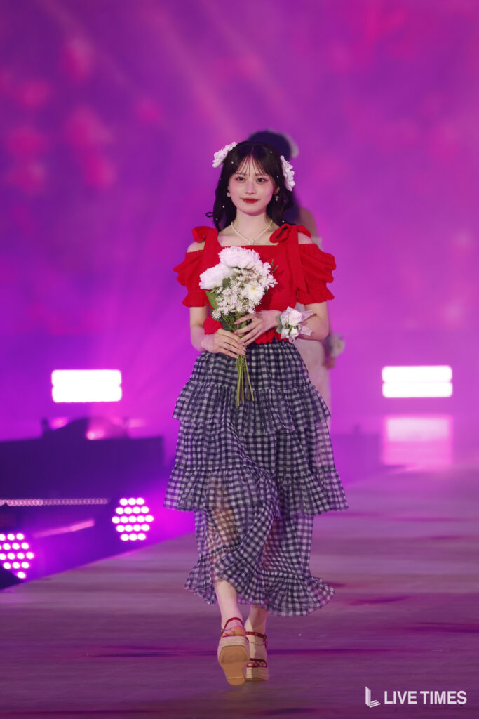 Young woman in a red off-shoulder top and tiered black-and-white gingham skirt walks on a pink-lit stage, holding a bouquet of white flowers.