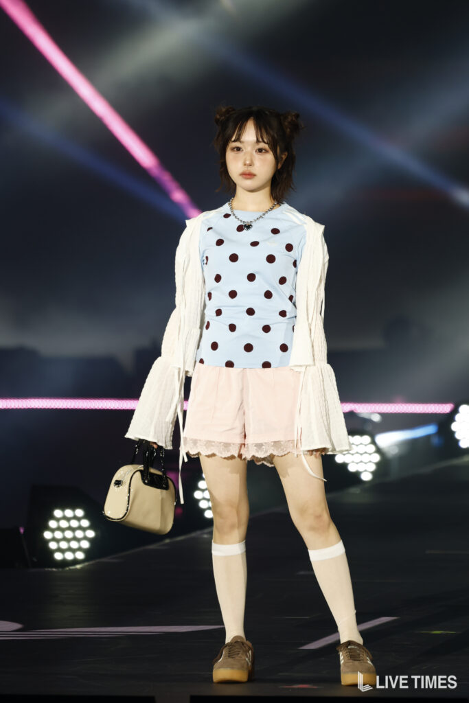 Young woman on a concert stage wearing a light blue polka-dot top, white cardigan, pink shorts, knee-high socks, and brown shoes, carrying a beige handbag.