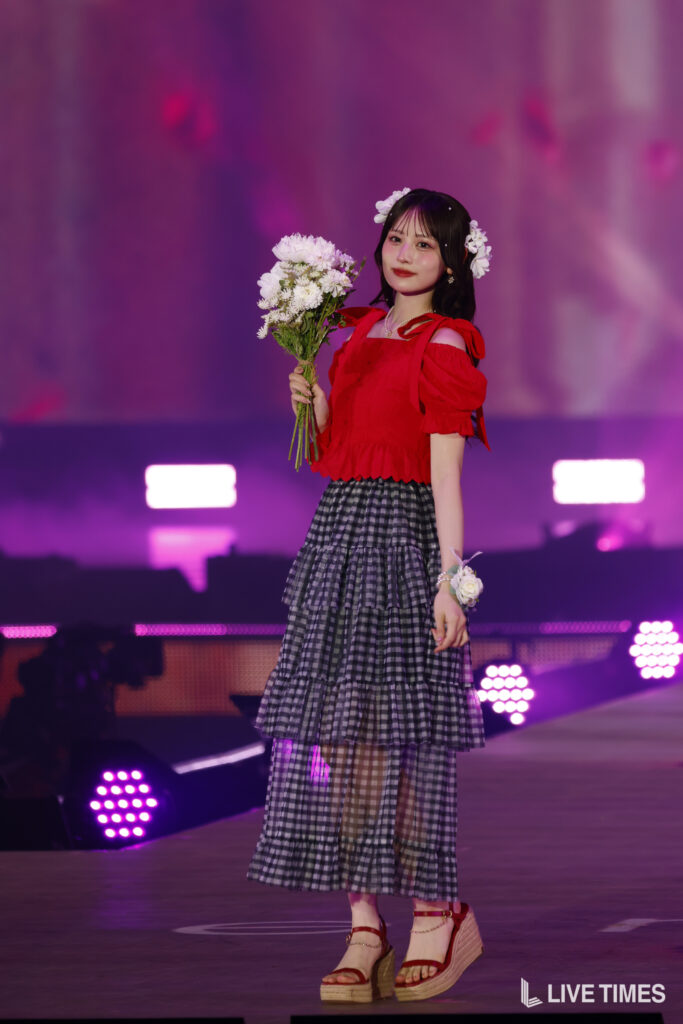 Young woman on a colorful stage holding a bouquet of white flowers, wearing a red off-shoulder top and a tiered plaid skirt with a flower crown.