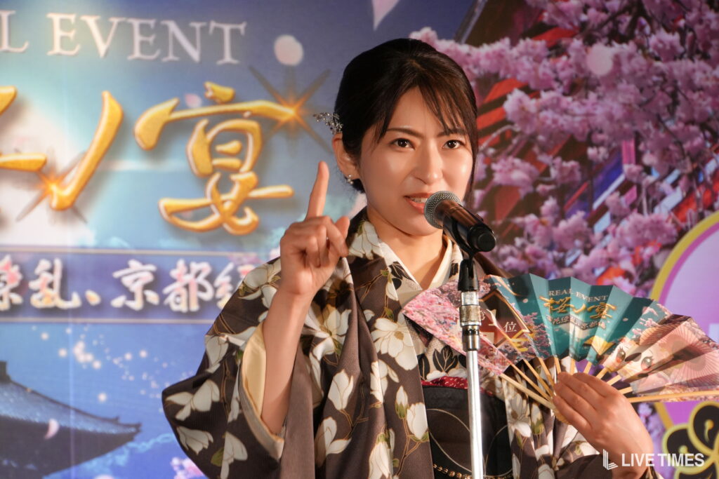 Woman in a floral kimono speaking into a microphone on stage, raising her index finger as she talks.