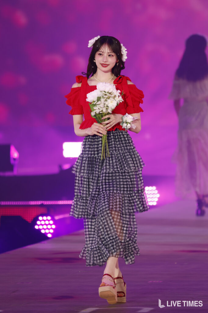 Young woman in a red off-shoulder top and tiered plaid skirt walks on a pink-lit stage, holding a bouquet of white flowers.