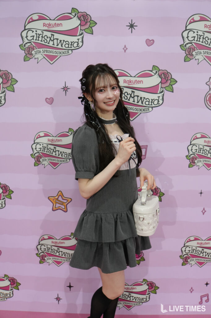 Young woman with long dark hair in a gray dress, posing and smiling at the camera with a white handbag at a pink GirlsAward backdrop.