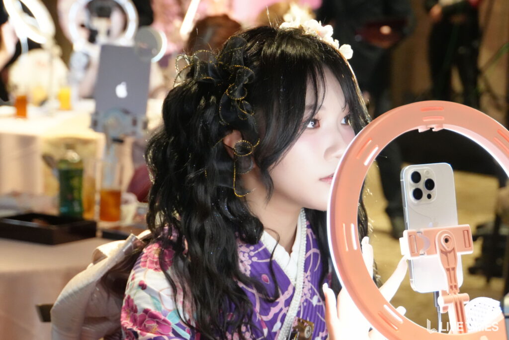 Young woman with black hair and a floral headpiece posing near a pink ring light and smartphone setup.