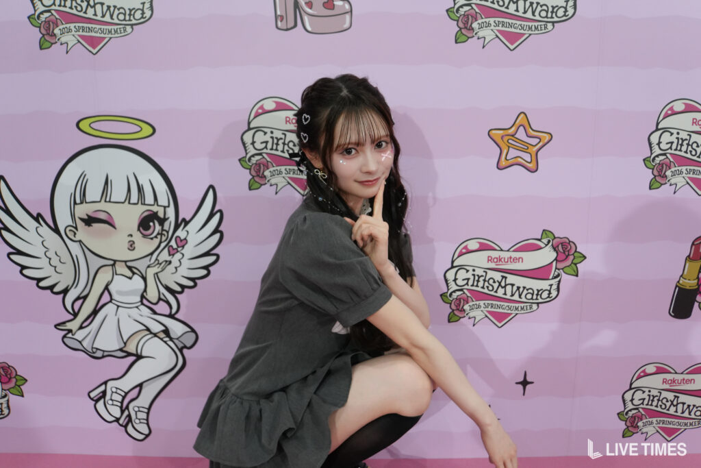 Young woman striking a pose against a pink backdrop decorated with GirlsAward logos and cartoon angels.
