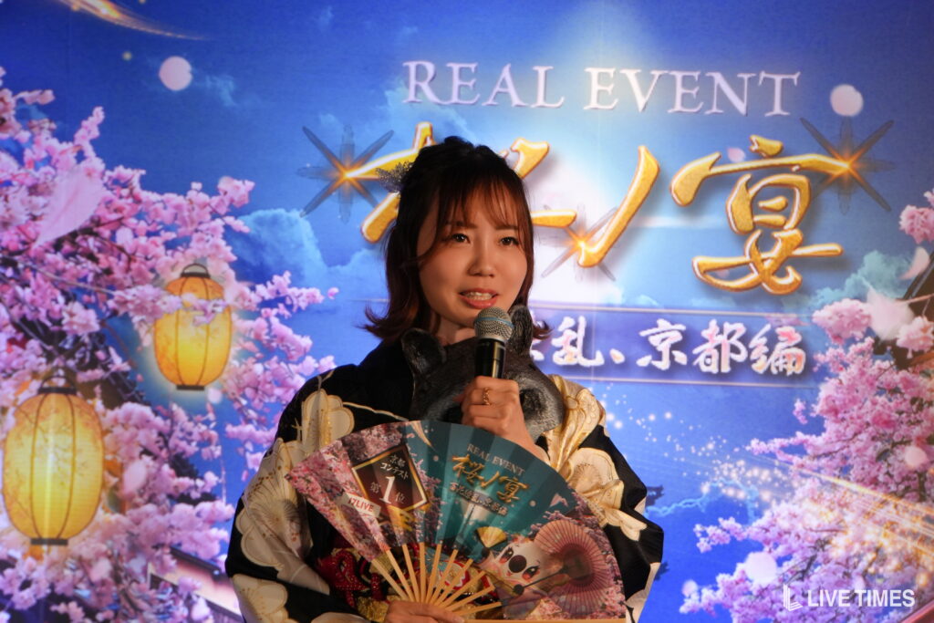 Woman in a traditional kimono speaks into a microphone on stage, with pink cherry blossoms and lanterns in a blue event backdrop behind her.