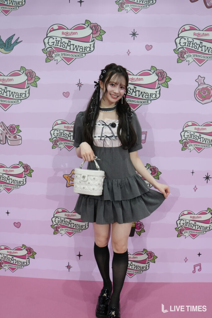 Young woman in a gray tiered dress posing on a pink backdrop with GirlsAward by Rakuten logos, holding a white handbag.