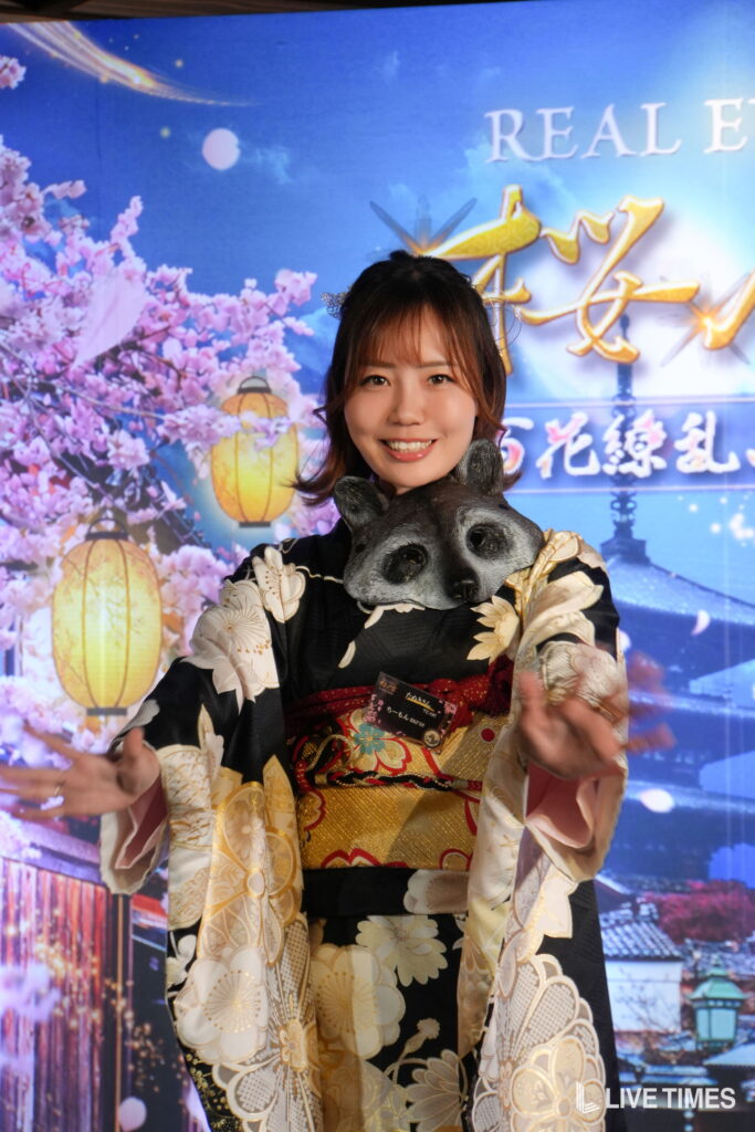 Woman in a floral kimono smiling, with a panda mask around her neck, posed in front of a blue festival backdrop with cherry blossoms and lanterns.
