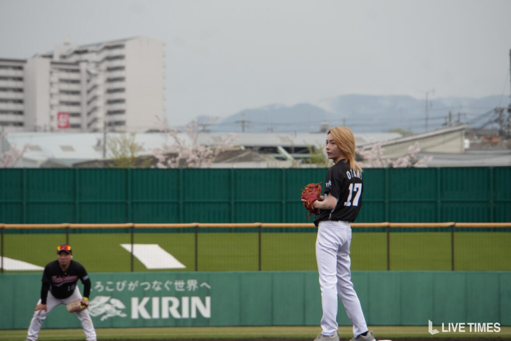 Baseball pitcher with blonde hair on mound during game, wearing black jersey and white pants, gloving left hand, infield and outfield fence behind him