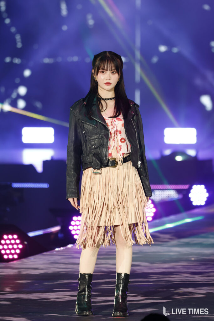 Female performer on a stage wearing a black leather jacket over a pink fringe dress and black boots; colorful concert lights in the background.
