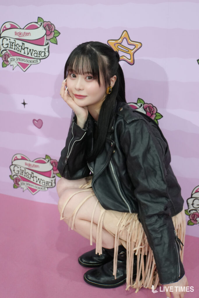 Young woman posing with her hand on her cheek in front of a pink GirlsAward backdrop, wearing a black leather jacket and beige fringe skirt.