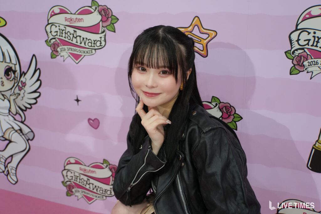 Young woman with black hair in a ponytail, wearing a black leather jacket, posing with a smile in front of a pink event backdrop.
