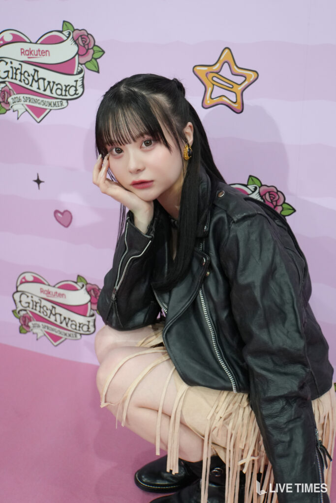 Young woman with long black hair wearing a black leather jacket, posing with her hand on her face against a pink backdrop with GirlsAward branding.