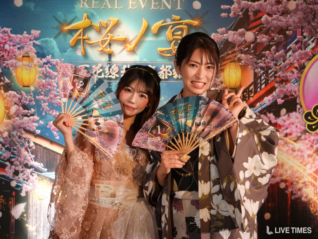 Two women in floral kimonos pose together, each holding decorative fans in front of a cherry blossom festival backdrop with lanterns and gold lettering.