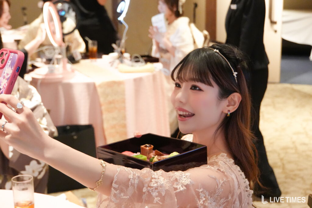 Young woman in a pink lace dress holds a bento box of sweets and poses for a selfie at a party with a pink phone in the foreground.