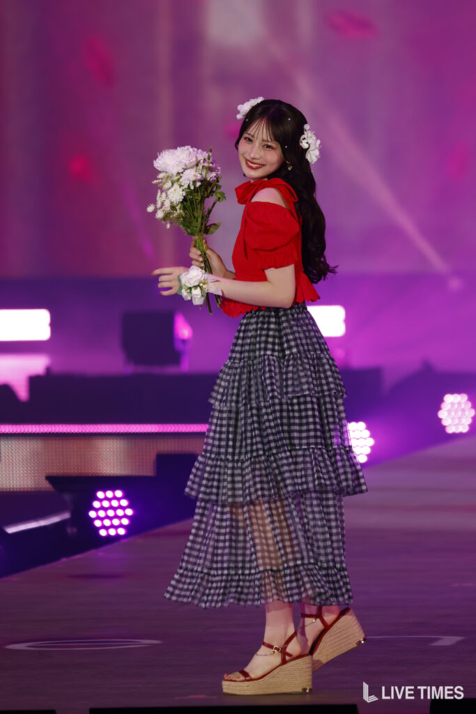 Young woman in a red off-shoulder top and tiered gingham skirt stands on a purple-lit stage, holding a bouquet of white flowers with floral hair accessories on both sides of her dark hair.