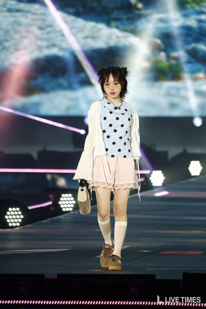 Young female performer walking on stage in a light blue polka-dot top, pink shorts, and a white cardigan, carrying a beige handbag.