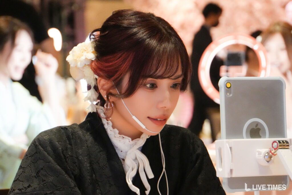 Woman with white flower hair accessory looking at a phone in a beauty booth, ring light in the background.