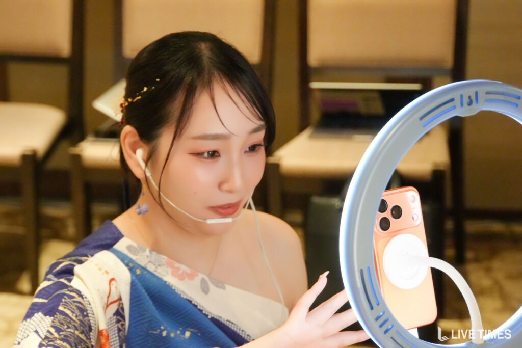 Young woman in a blue patterned kimono looks at a pink device mounted on a blue ring light, wearing a white face mask under her chin.