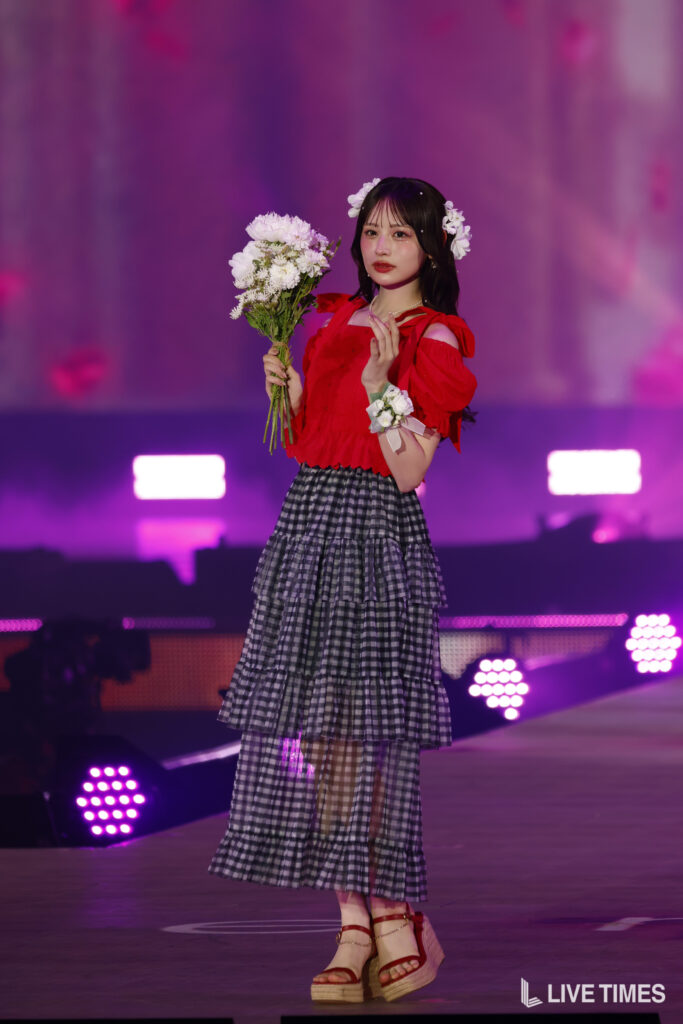 Female performer on stage in a red off-shoulder blouse and tiered plaid skirt, holding a bouquet of white flowers with a flower crown, pink stage lights in the background