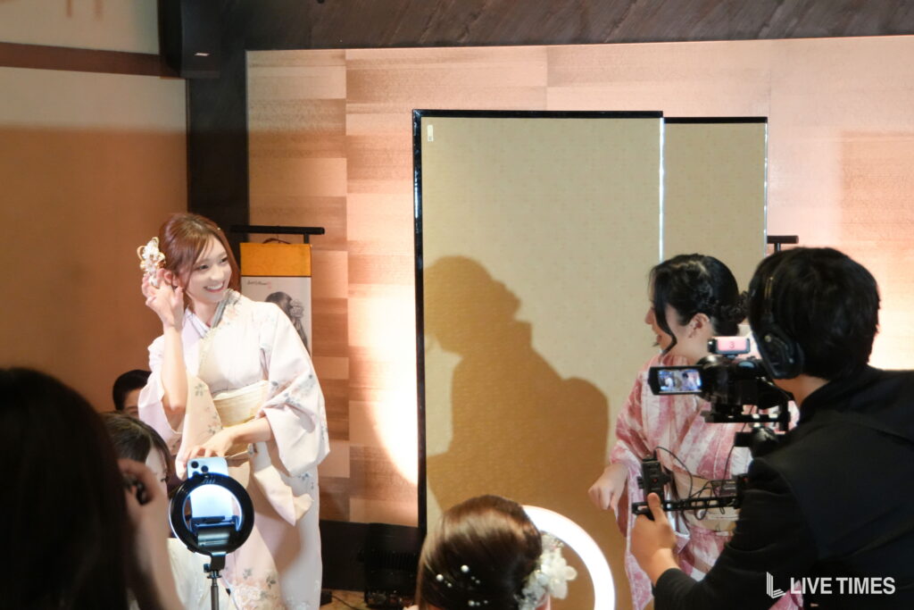 Young woman in a pale kimono smiles as photographers film at a fashion event, a bright ring light in the foreground.