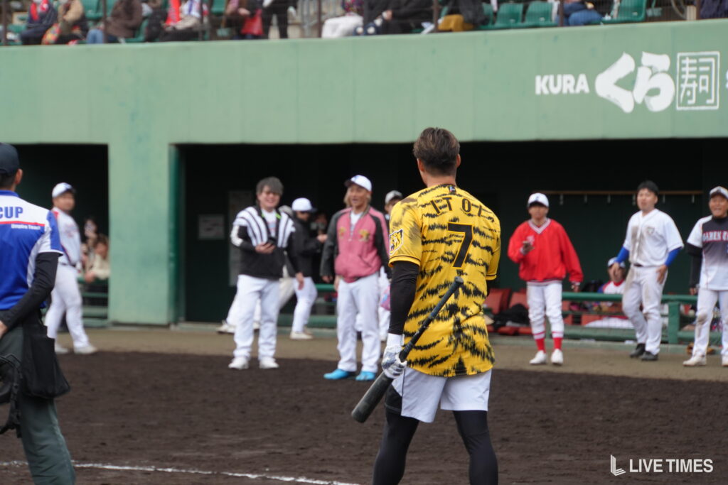 Baseball player in a yellow tiger-striped jersey with number 7 stands at home plate holding a bat, facing teammates in the dugout.