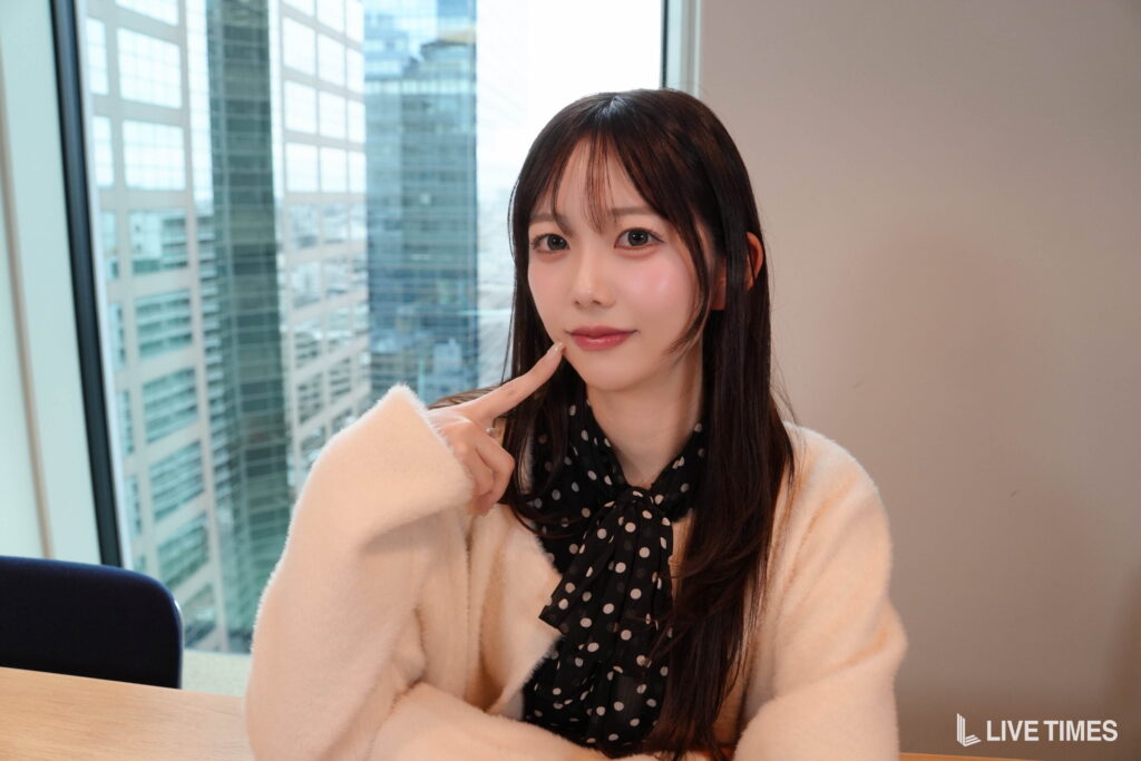 Woman posing indoors with a polka-dot scarf and fluffy beige coat, touching her chin as city buildings are visible through a window behind her, watermark LIVE TIMES.