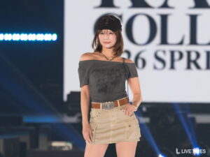 〈インタビューあり〉人気ライバー・えりか、オフショル×ミニスカで美スタイル披露。ストリートコーデで関コレのランウェイ彩る【KANSAI COLLECTION 2026 S/S】 Young woman posing on stage in an off-shoulder gray top, beige cargo skirt, belt, and cap; spotlight and large backdrop behind her.