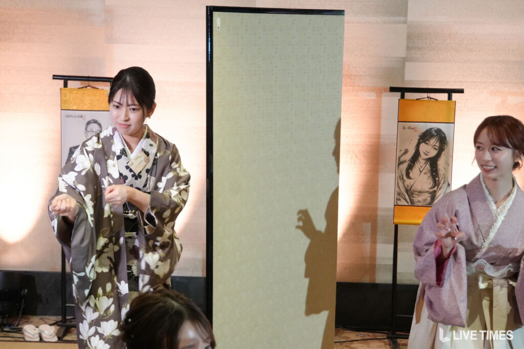 Two women in traditional kimonos perform a dance on a stage, with decorative screens behind them and posters visible in the background.