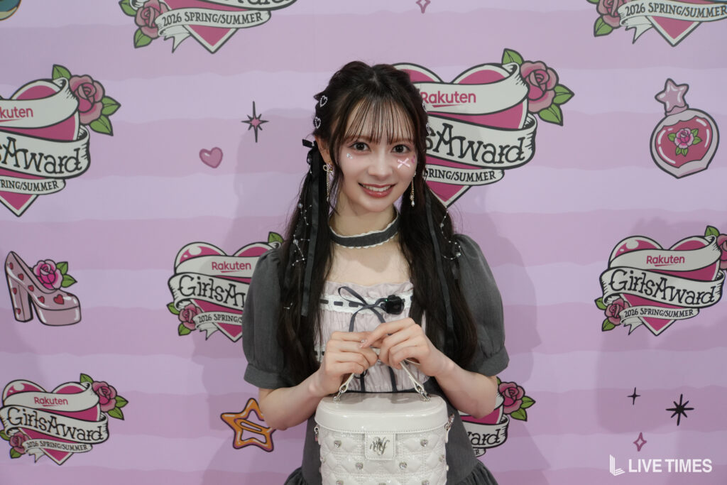Young woman posing in front of a pink GirlsAward backdrop with Rakuten logos, holding a white handbag.
