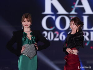 Two women in evening gowns pose on a dark stage, smiling at the camera with a large blurred logo in the background (LIVE TIMES).