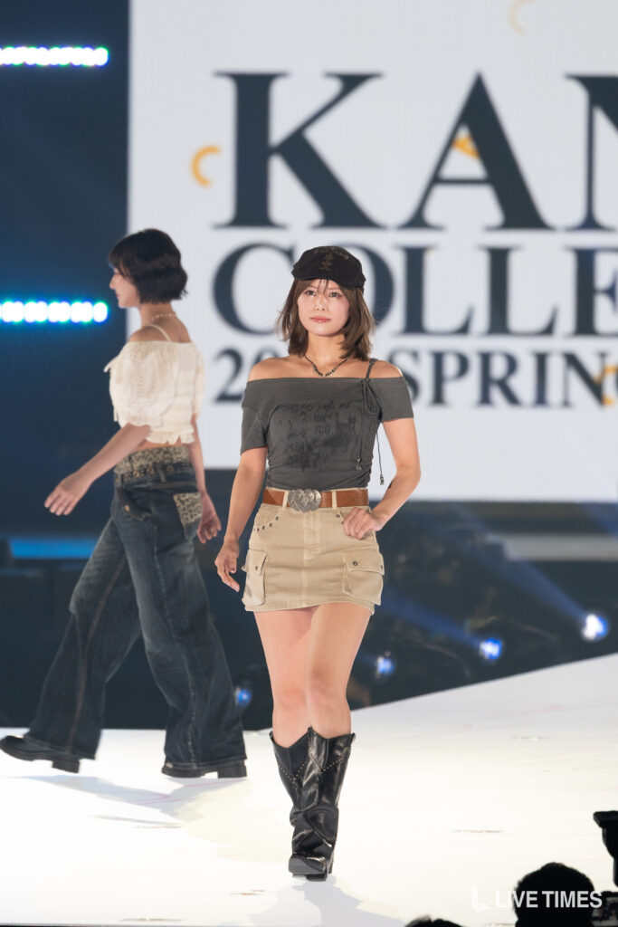 Front-facing model walking the runway in an off-shoulder gray top, beige cargo skirt, black hat, and knee-high boots; large stage screen behind shows branding.