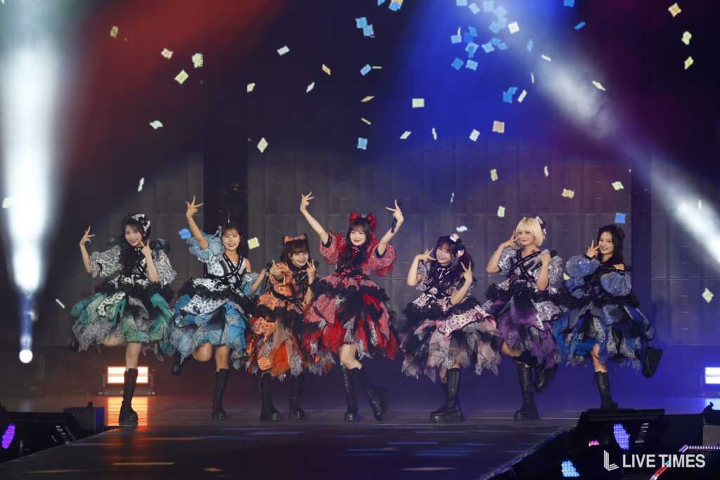 Eight performers in vivid, layered dresses dance on a lit stage as confetti falls around them.