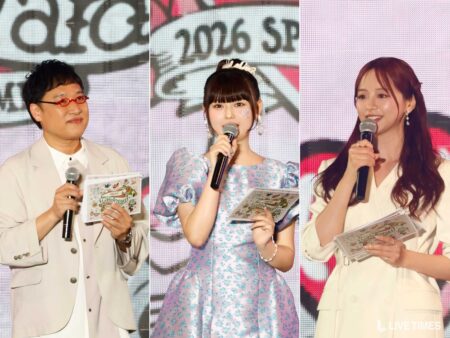 Three hosts on a stage hold microphones and decorative booklets; left is a man with light suit and red glasses, center a girl in a blue floral dress with a tiara, right a woman in white blazer.