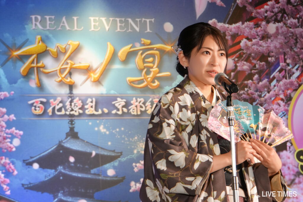 Woman in a floral kimono speaks into a microphone on stage, with pink cherry blossoms backdrop and blue sky imagery behind her.