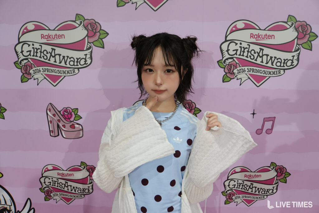 Young woman posing in front of a pink backdrop with GirlsAward logos, wearing a light blue polka-dot dress and a white shawl.