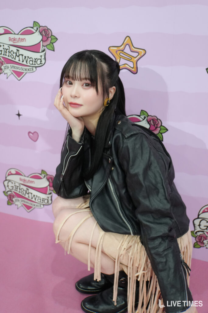 Young woman posing in a black leather jacket and beige fringe skirt against a pink backdrop with event logo on the wall.