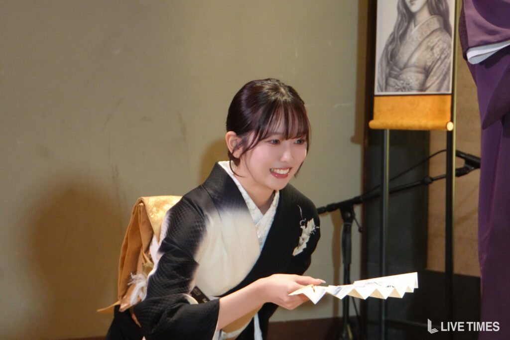 Young woman in a black and cream kimono smiling while handling folded paper fans on stage. (performer)