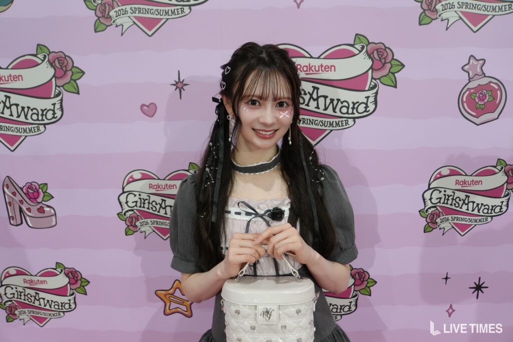 Young woman posing at a GirlsAward event, pink backdrop with logos, wearing a gray outfit and holding a white handbag.
