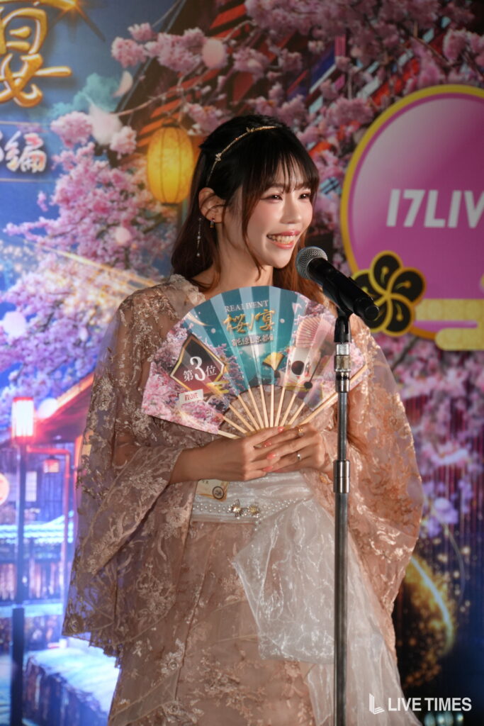 Smiling woman in a pink lace dress holding a decorative fan at a microphone on stage.