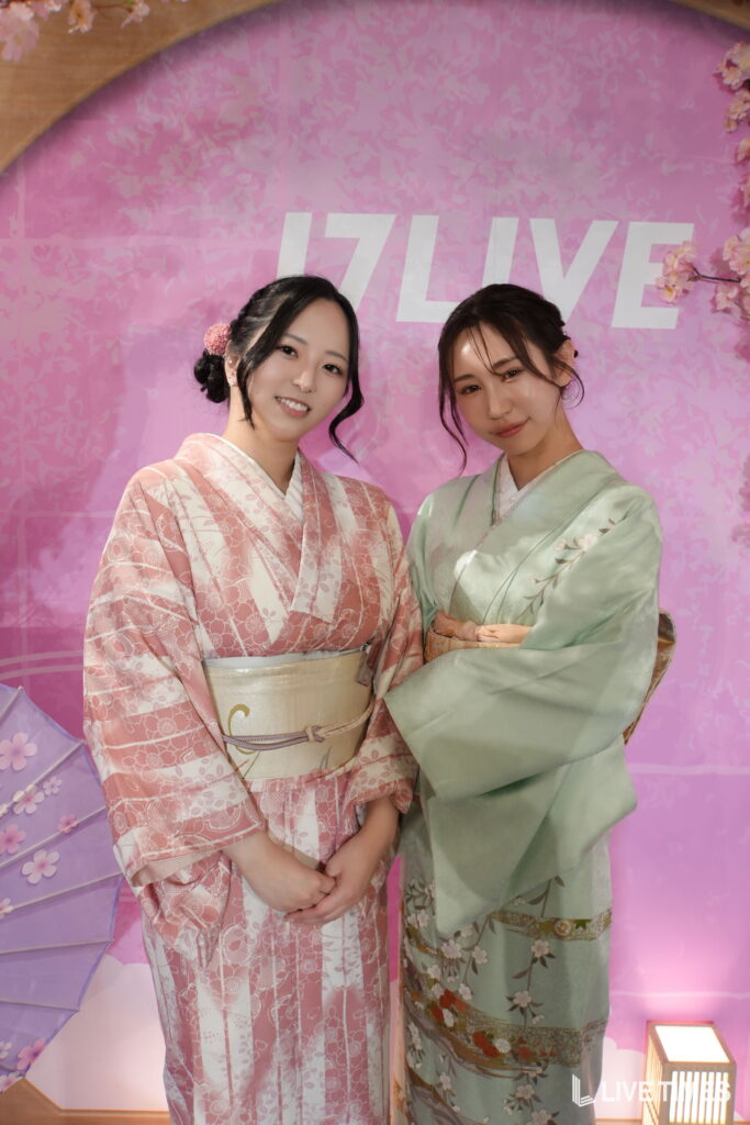 Two women in pastel-colored kimonos posing together in front of a pink floral backdrop, smiling at the camera.