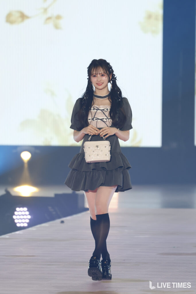Runway model in a dark frilly dress walks forward, holding a small pale handbag with a bright stage backdrop behind her.